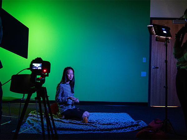 A student sits on a blanket on the floor in a darkened room with a green screen behind. A camera stands in the foreground, recording the scene.
